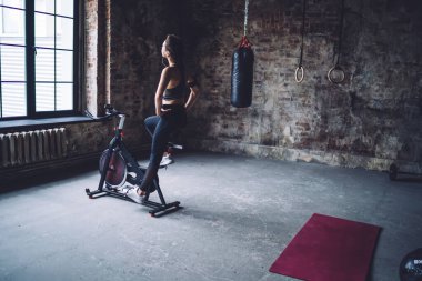 Arka plan ciddi beyaz kadın spor salonunda modern bisiklet simülatöründe egzersiz yapıyor. Zayıflatıyor ve formda kalıyor. Kulaklıklı kadın, kilo vermek ve antrenman yapmak için zaman harcıyor.