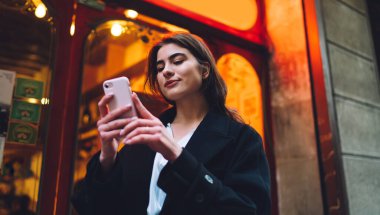 Yoğunlaştırılmış gülümseyen, günlük kıyafetlerle cep telefonuyla gezen bir bayanın akşamları bina yakınında yürürken durduğu bir yerden.