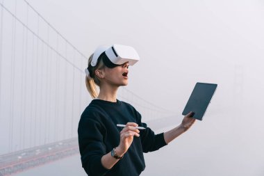 San Francisco 'da Golden Gate yakınlarında dikilirken dijital kalem ve tablet kullanan VR gözlüklü genç bir kadının yan görüntüsü