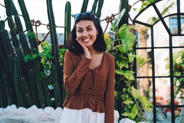 Positive smiling young lady wearing trendy dress standing in green park against big cactuses while touching cheek coquettishly and looking at camera