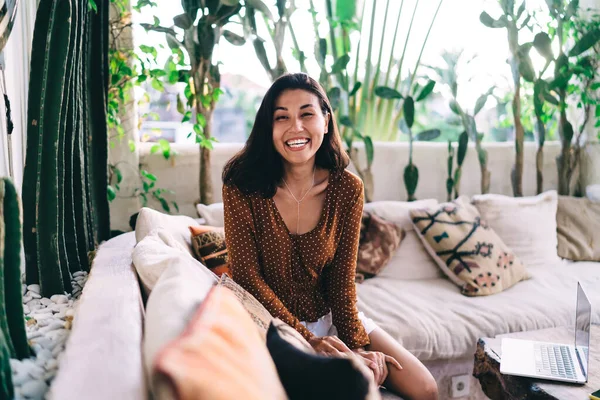 Positive young female in casual outfit smiling at camera while having rest on comfortable sofa in cozy light living room