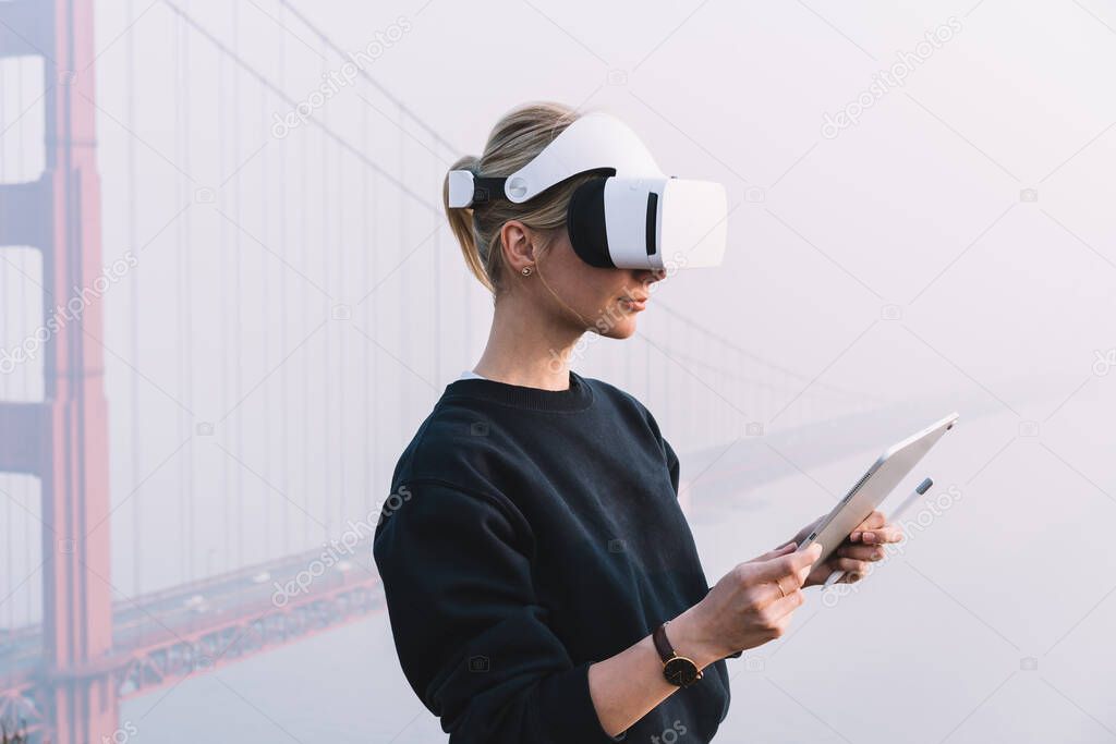 Side view of concentrated female in stylish clothes standing against bright bridge while wearing headset and holding digital tablet with pen