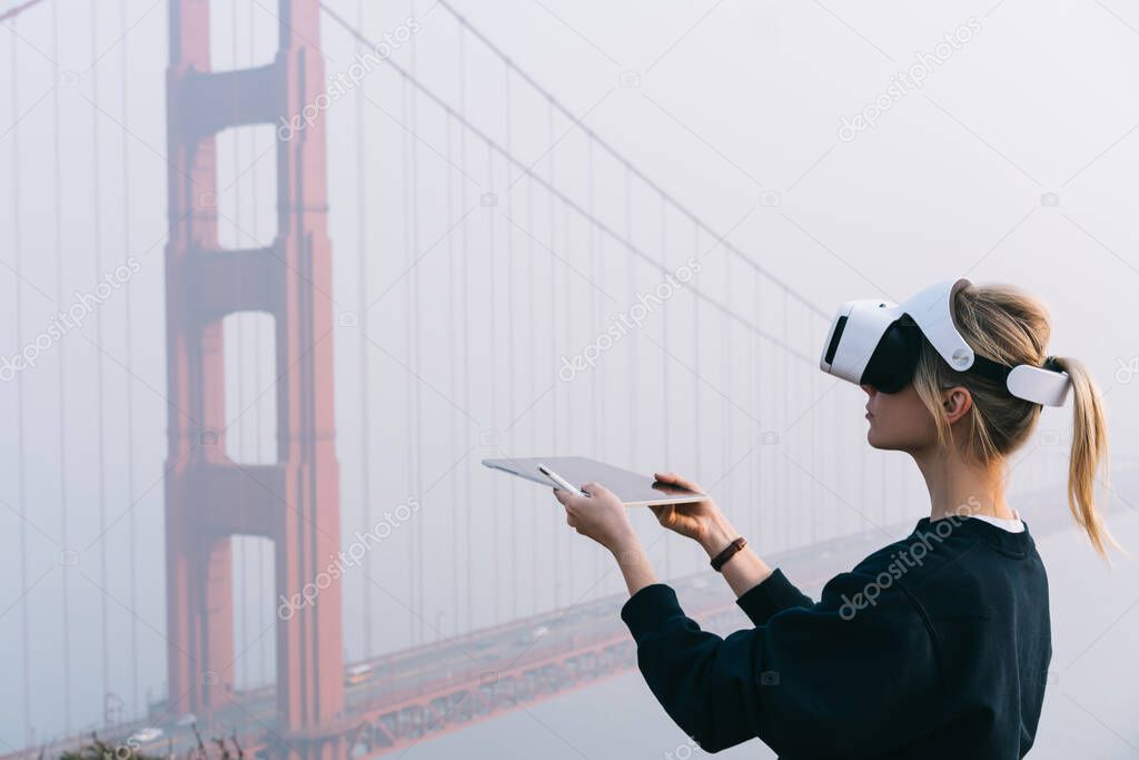 Side view of female with short blond hair in black sweater holding tablet and pencil while standing near bridge located in San Francisco and wearing VR headset