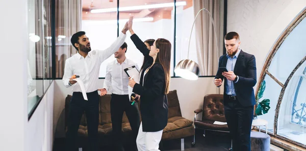 stock image Group of positive multiethnic colleagues in formal clothes standing in modern office and giving high five to each other while celebrating triumph