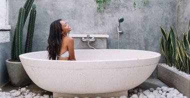 Mutlu Asyalı kadın egzotik tatil beldesinde banyo yapıyor, eğleniyor ve modern tasarımın tadını çıkarıyor, gülümseyen kadın beyaz küvette oturuyor ve tatilde vücut derisi ve hijyenine önem veriyor.