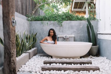 Beyaz bikinili çekici bir kadın Bali 'deki kaplıca merkezinde lüks bir küvette dinleniyor ve kameraya bakıyor.