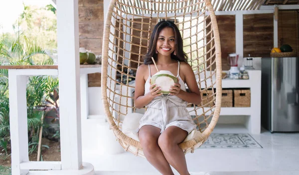 Cheerful ethnic female in casual clothes smiling and sitting on wicker hanging egg chair while drinking coconut during rest on Bali