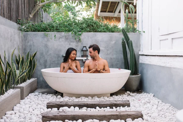 stock image Funny multiracial male and female couple in love sitting in white bathtub in namaste pose recreating, just married young family spending honeymoon on villa enjoying water and relaxation in tub