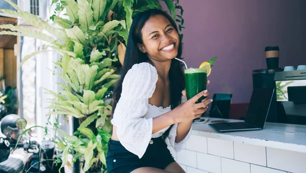 穿着夏装的乐观的年轻女性微笑着看着镜头 一边在热带酒吧里享受着健康的绿色鸡尾酒 — 图库照片