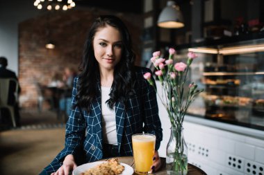 Zarif giysiler içinde gülümseyen bir kadın ahşap masada oturuyor ve rahat bir kafede kameraya bakarken lezzetli bir kahvaltı yapıyor.