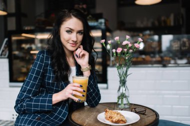 Resmi giysiler içinde neşeli bir kadın yuvarlak masada oturuyor ve sabah kafede kahvaltı ederken taze meyve suyu içiyor.