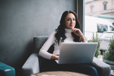 Masanın yanında kucağında bilgisayarla oturan ve çağdaş ofiste uzaktan proje üzerinde çalışan içerikli kadın kameraya bakıyor