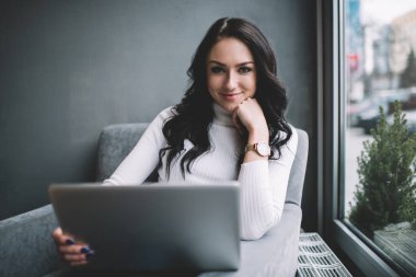 Kıvırcık siyah saçlı neşeli bir kadın işçi, modaya uygun balıkçı yaka kazaklı. Bilgisayardan finansal haberler hakkında mesajlar okuyor, kameraya bakıyor.