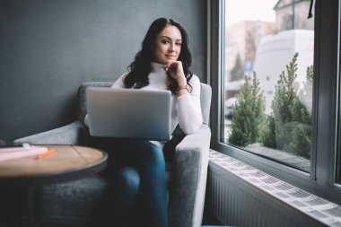 Düşünceli kadın serbest çalışan, pencerenin yanında otururken kameraya sırıtarak bakıyor ve gündüzleri yeni iş planı üzerinde çalışıyor.
