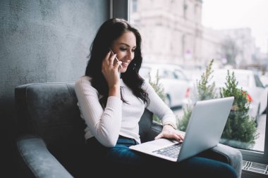 İyimser kadın, beyaz kazaklı, rahat koltukta dinlenirken ve cep telefonuyla sohbet ederken gülümseyen ve bilgisayarını karıştıran serbest yazar.