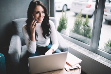 Dizüstü bilgisayar kullanan ve cep telefonuyla konuşan mutlu kadın serbest yazar yaratıcı iş yerinde oturup iş projesinde çalışıyor.