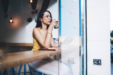 Gözlük takmış, gazete okurken kahve içen ve pencereden dışarı dalgın dalgın bakan bir kadın bardağının içinden.