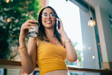 Gözlüklü, yaz tişörtlü, tek kullanımlık kahve bardağı tutan, geniş pencereli ve gözlerini kaçıran konuşkan genç bir kadın.