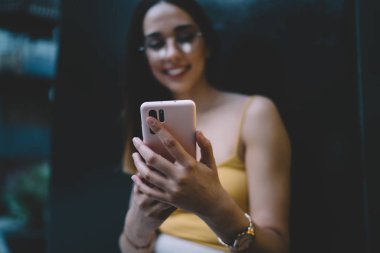 Sıradan kıyafetler ve gözlüklerle gezen neşeli genç bir kadın akıllı telefondan selfie çekiyor. Şehir binasının siyah duvarının yanında dururken mobil parmak izi tarayıcısı kullanıyor.