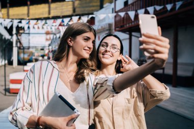 Neşeli Kafkasyalı hippi kızlar günlük giysiler giyip kentteki dostça buluşmaları hatırlamak için özçekim fotoğrafları çekiyorlar. Neşeli kadın blogcular modern akıllı telefon kamerasıyla video bloğu çekiyorlar.