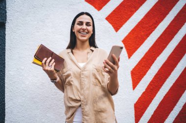 Akıllı telefonlara bakarken kameraya gülümseyen ve şehir caddesinde şık bir duvarın arkasında not defterleri tutan yaratıcı genç bayan.