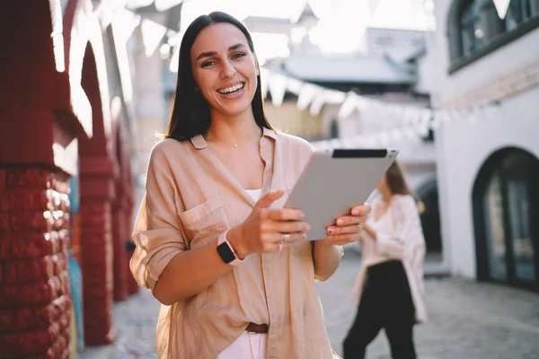 Young smiling female remote worker in beige blouse with smartwatch standing on pavement with tablet and laughing while looking at camera