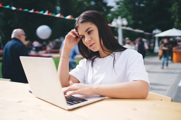 阳光明媚的日子里 身穿白衬衫的年轻黑发女子坐在公园露天咖啡馆的桌子边 一边浏览笔记本电脑 一边看屏幕 — 图库照片