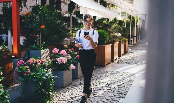 身穿休闲装和眼镜 背着背包的积极的年轻女子手持智能手机和平板电脑走在花店附近 — 图库照片