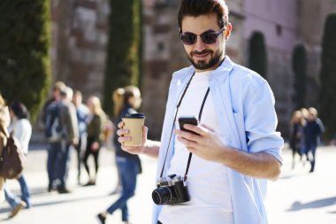 Düşünceli, sakallı, günlük kıyafetler ve güneş gözlüklü, elinde fotoğraf makinesi, elinde kahve fincanı ve akıllı telefonla sörf yapan bir adam.