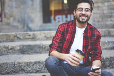 Kırpılmış, sakallı, kareli tişörtlü ve gözlüklü mutlu bir erkek cep telefonuyla mesajlaşırken ve taştan merdivenlerde kahve içerek boş zamanların tadını çıkarıyor.