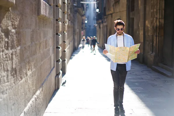 Full body young male in casual outfit and eyeglasses looking at map and checking route while strolling on street in old town