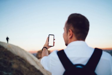 Beyaz elbiseli, siyah sırt çantalı, kayanın yanında selfie çekmek için akıllı telefon kullanan kimliği belirsiz bir adamın arka planı.