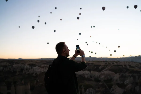 观看热气球飞越高山时 带背包的年轻人用手机拍照的侧视图 — 图库照片
