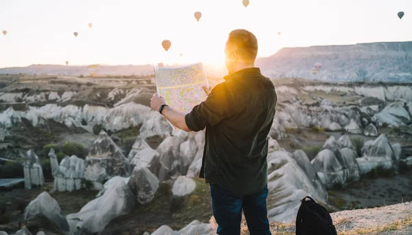 背对着地图的匿名男性游客背对着地图 站在山上 背对着岩石地形上的热气球 — 图库照片