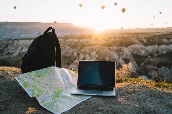 Laptop on map with backpack on hill with sunrise on horizon with rocky terrain and hot air balloons in background