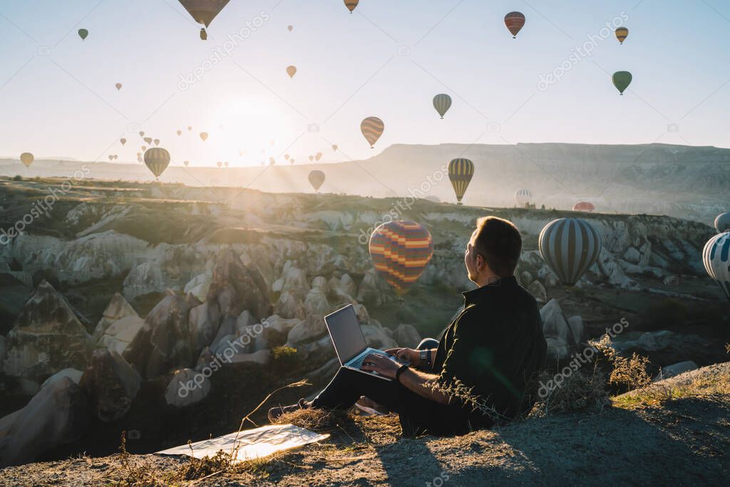Back view of man using laptop and looking at colorful hot air balloons while sitting on ground near rocky terrain in back lit