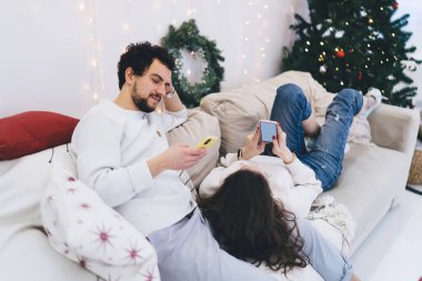 Beyaz erkek milenyumu, kış tatillerinde elinde cep telefonuyla şenlikli bir dairede kanepeye uzanmış kız arkadaşı cep telefonuyla e-posta gönderirken yakışıklı bir adam kameraya bakıyor.