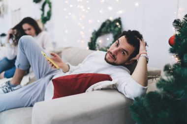 Beyaz erkek milenyumu, kış tatillerinde elinde cep telefonuyla şenlikli bir dairede kanepeye uzanmış kız arkadaşı cep telefonuyla e-posta gönderirken yakışıklı bir adam kameraya bakıyor.
