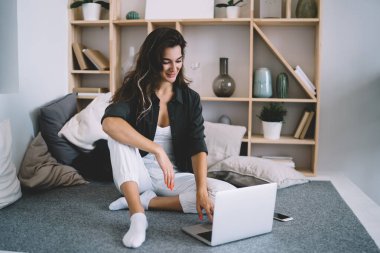 Kanepede oturup dizüstü bilgisayarı karıştıran ve karantinada zaman geçiren neşeli, genç bir kadın.