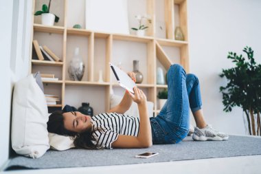 Kot pantolon ve çizgili tişört giyen, tabletleri karıştıran, ayakları çaprazlanmış, rafların yanında dinlenen tüm vücuda bakan genç kadın.