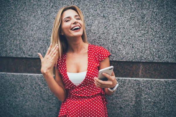Happy Caucasian girl in polka dots sundress enjoying mobile networking at publicity area satisfied with roaming tariffs, prosperous female blogger satisfied with received invitation message