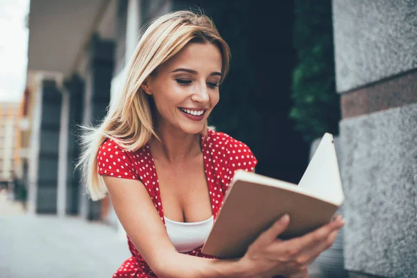 Sincerely female enjoying recreation time for reading favourite literature best seller during leisure time in urban city, happy Caucasian woman 20 years old interested on positive plot of novel book