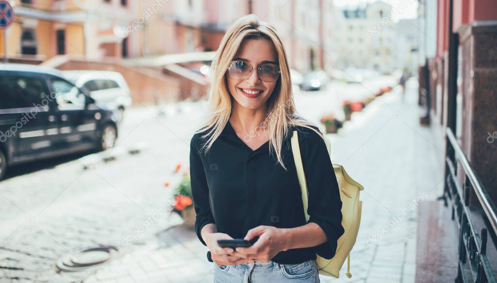 Half length portrait of carefree female tourist in trendy glasses for sun protection walking in town during summer recreation vacations, millennial traveller with modern smartphone gadget posing