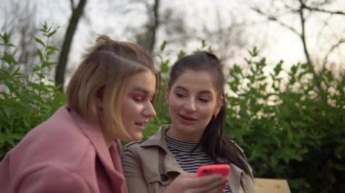 Kadın arkadaşlar telefon ekranına bakıyor, parkta yürüyor, arkadaşlık kavramı.