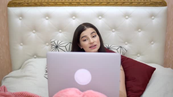 Joven mujer sonriente hablando por chat de vídeo en la cama usando la conversación de vídeo portátil — Vídeo de stock