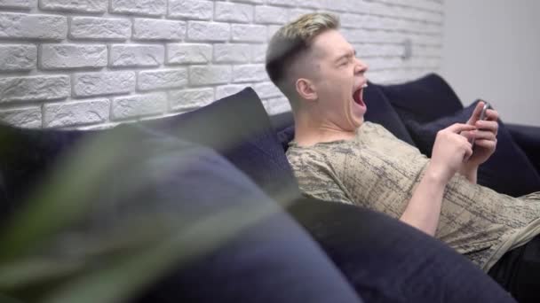 Oung faul gelangweilt zu Hause Mann gähnen, entspannen auf dem Sofa, mit Handy — Stockvideo