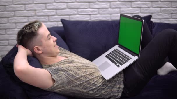 Glücklicher junger Mann entspannt sich auf dem Sofa, sucht Laptop-Green-Screen-Attrappe, gemütliches Zuhause — Stockvideo