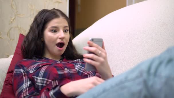 Молодая привлекательная шокированная женщина на диване говорит WOW с помощью мобильного телефона, уютный дом — стоковое видео