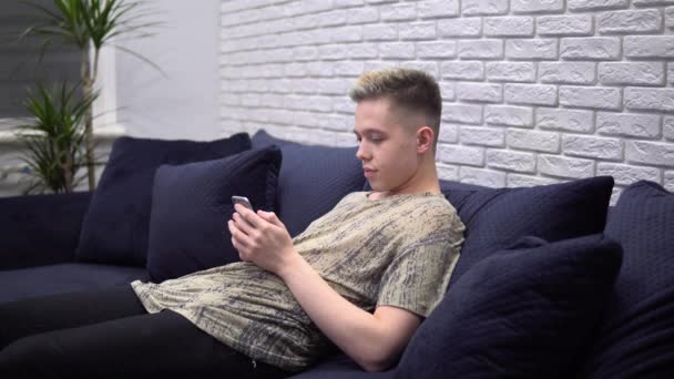 Молодой человек использует смартфон, серфинг Интернет, расслабляясь на диване дома — стоковое видео