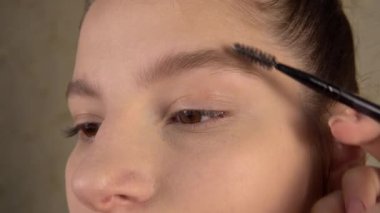 Makyaj sanatçısı kaşlarını fırçalıyor, model müşteri yüzünü kapatıyor.
